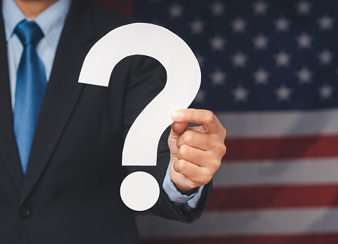 Question mark symbol. Businessman in a suit holding a white question mark paper while standing on the American flag background. Close-up photo. Space for text