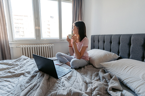 Beautiful young woman drinking coffee while working on the laptop from her bed.