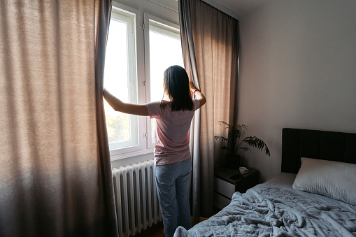 Beautiful young woman opening up curtains after waking up in the bed in the morning.