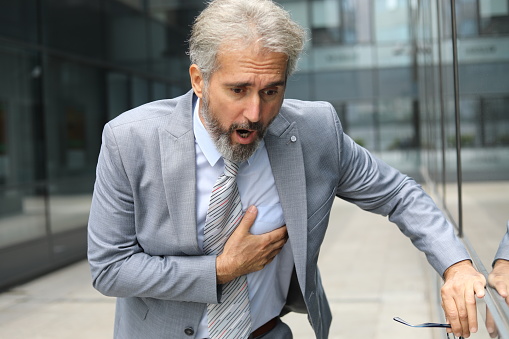 Pre-infarction condition. A senior man in a suit stands on the street near an office center holding his heart. He feels a sharp pain in his chest. He needs urgent medical help.