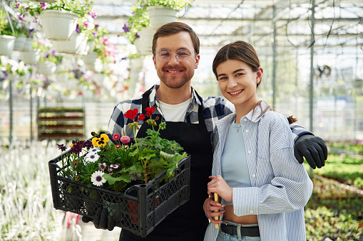 Sincere smile, standing, holding flowers. Man and woman are together in bright greenhouse.