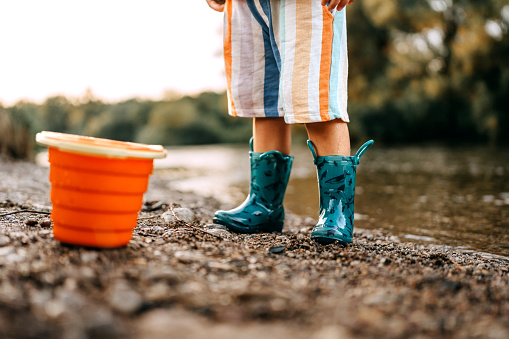 Close up shot of an unrecognisable little boy wearing rubber boots standing by the river with orange bucket toy