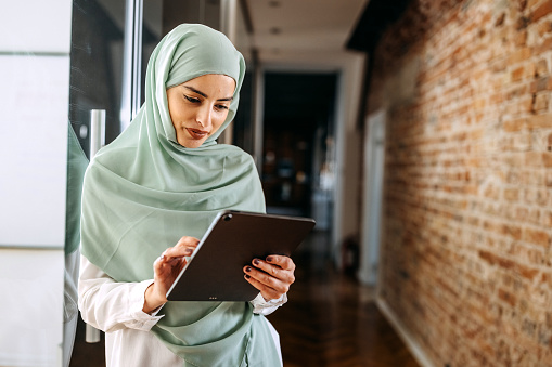 Young beautiful woman wearing hijab using digital tablet at modern office hallway