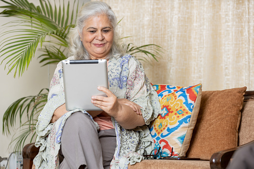Beautiful Indian senior woman in grey hair using tablet and smiling while relaxing on the couch at home