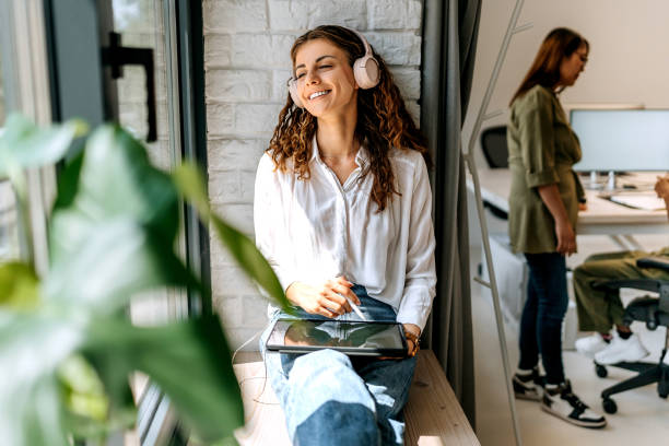 istock Young beautiful woman with headphones using digital tablet while sitting by the window at modern office 1663293393