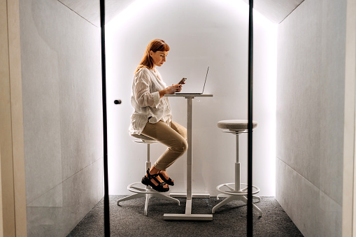 Young pregnant businesswoman using phone while working in an office pod