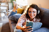 Woman using mobile phone and listening to music on wireless headphones, while relaxing on the sofa