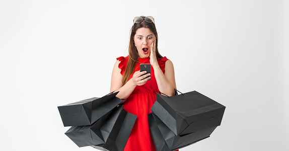 Surprised woman doing online shopping on Black Friday, she is holding a smartphone and many shopping bags
