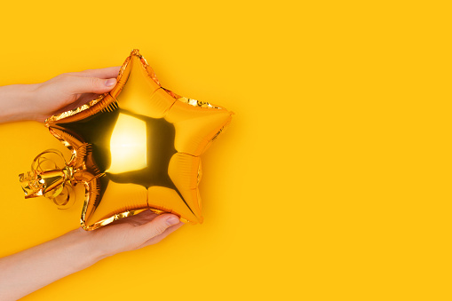 Female hands hold gold colored inflatable foil balloon in a star shape. Creative concept on a yellow background.