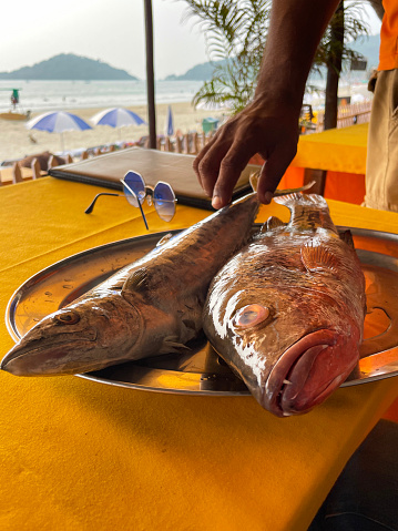 Stock photo showing close-up view of outdoor seating and tables of a restaurant on beach with metal serving platter of freshly caught kingfish and red snapper selected by restaurant patrons to be cooked and served for a meal.