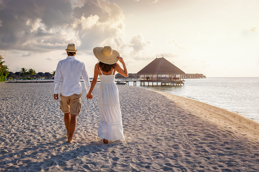 A beautiful tourist couple in white summer clothing walks along a tropical beach in the Maldives holding hands during sunset time
