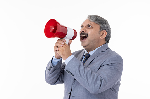 Angry senior male professional dressed in suit screaming into megaphone while standing against white background