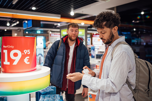 Couple wearing casual clothing exploring a duty free shop in an airport in Germany before catching their flight. One of the men is looking at aftershave, spraying it onto his wrist.