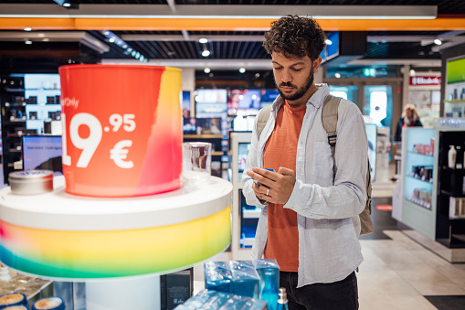 Man wearing casual clothing exploring a duty free shop in an airport in Germany before catching his flight. The man is looking at aftershave.