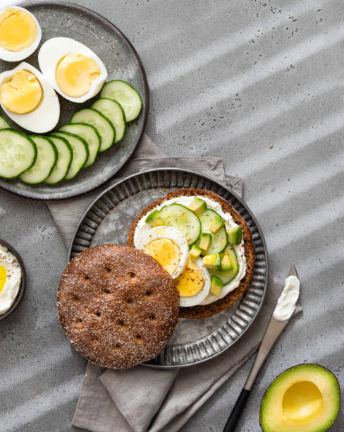 istock Rye bread buns for cooking sandwiches with cream cheese spread, boiled eggs, avocado cubes and sliced fresh cucumber. Top view, grey background. 1663429363