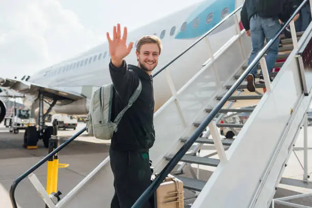 A young backpacker getting onto a plane while budget travelling. He is walking up the steps to the plane carrying his suitcase, smiling at the camera while waving.
