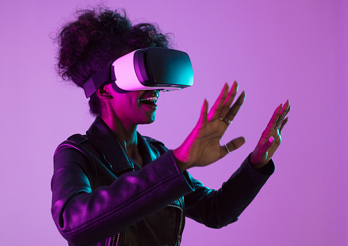 Side view of excited afro american woman wearing leather jacket and VR glasses posing over bright vivid pink and purple ultraviolet background, raising hands.