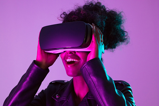 Excited afro american woman wearing leather jacket and VR glasses posing over bright vivid pink and purple ultraviolet background, looking away and smiling.