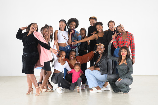 A group of beautiful black females consisting of toddlers, teenagers and young woman together in a group shot in a studio. They are Laughing and joking while they are looking at the camera.