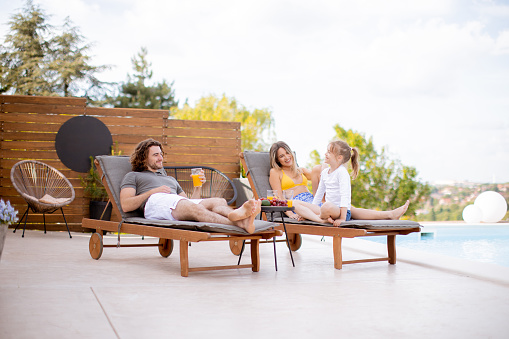 Happy family with a mother, father and daughter sitting on the deck chairs by the swimming pool