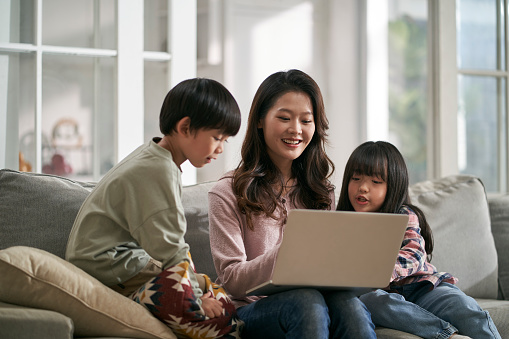 young asian mother and two children sitting on family couch at home using laptop computer together