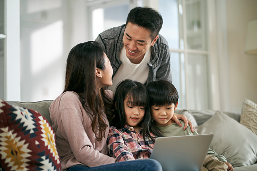 happy young asian couple with two children sitting on family couch at home using laptop computer together