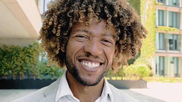 Close-up video of a handsome charismatic confident curly haired brazilian or hispanic man, successful entrepreneur, standing outdoors, looking at the camera, smiling friendly