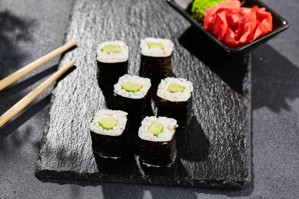 istock Avocado hosomaki on dark stone table. Hosomaki with avocado. Sushi roll with avocado inside. Style concept japanese menu with black background, leaves and hard shadow. 1663571328