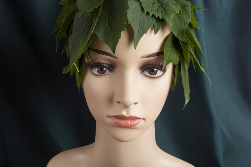 a display mannequin wearing hair of leaves and representing a nature goddess in front of a green curtain. Minimal color still life photography.