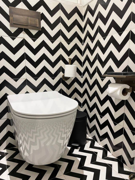 Close-up image of luxury hotel bathroom, black and white zig-zag patterned wall and floor tiles, wall hung ceramic toilet, brass button flush, phone holder shelf, toilet roll holder and paper, pedal bin Stock photo showing a hotel bathroom featuring a white toilet with modern fittings include bathroom hygiene in the form of a hand shower bidet head attachment, which has been fixed to the wall next to the WC, complete with wall bracket and brass hose connection. pedal bin stock pictures, royalty-free photos & images