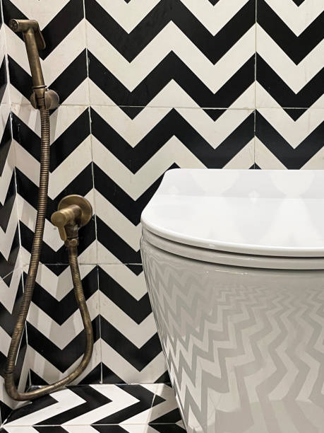 Full frame image of luxury hotel bathroom with black and white zig-zag patterned wall tiles, wall hung ceramic toilet, brass hand shower bidet head attachment Stock photo showing a hotel bathroom featuring a white toilet with modern fittings include bathroom hygiene in the form of a hand shower bidet head attachment, which has been fixed to the wall next to the WC, complete with wall bracket and brass hose connection. pedal bin stock pictures, royalty-free photos & images