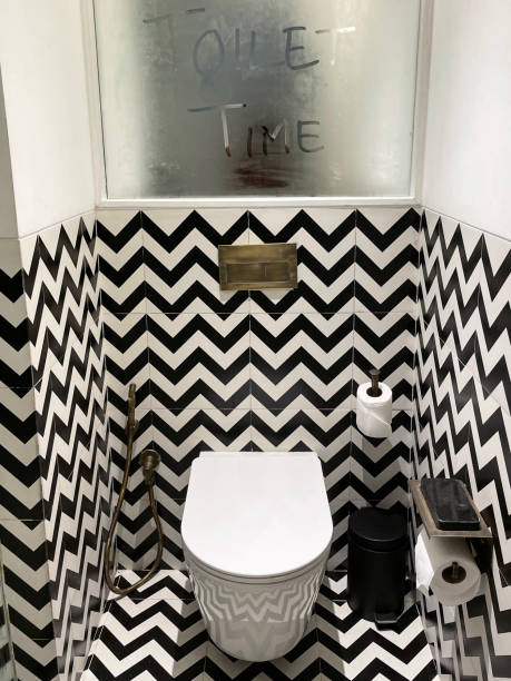 Image of luxury hotel bathroom, black and white zig-zag patterned wall and floor tiles, steamed up mirror, wall hung ceramic toilet, brass hand shower bidet head attachment, phone holder shelf, toilet roll holder and paper, pedal bin Stock photo showing a hotel bathroom featuring a white toilet with modern fittings include bathroom hygiene in the form of a hand shower bidet head attachment, which has been fixed to the wall next to the WC, complete with wall bracket and brass hose connection. pedal bin stock pictures, royalty-free photos & images