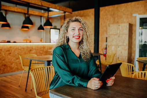 Cheerful businesswoman with curly hair sitting at table holding digital tablet and smiling at camera in modern office breakout space