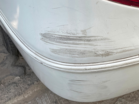 Stock photo showing close-up view of a white car rear bumper that has been freshly scratched after a parking accident. This photo concept shows primer and colour coat scratches in need of respraying at a garage, and maybe a car insurance claim.