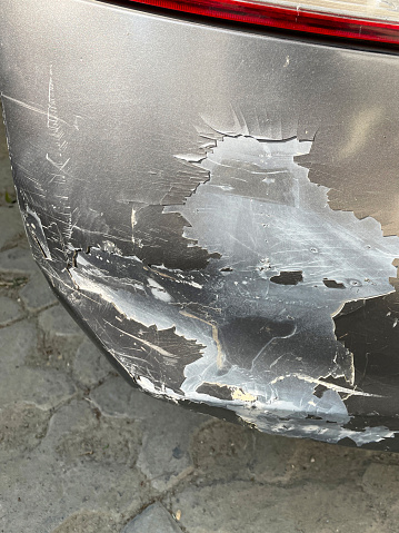 Stock photo showing close-up view of a silver car front bumper that has been dented and scratched after a parking accident. This photo concept shows primer and colour coat scratches in need of respraying at a garage, and maybe a car insurance claim.
