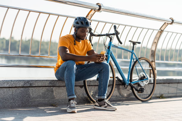 Happy multiracial man wearing protective helmet sitting at the street and holding mobile phone