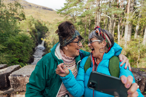 Waist-up of two senior female friends in the middle of hiking up the mountain landscape surrounding Loch Torridon in the Northwest Highlands, Scotland. They are side by side taking a selfie on a smartphone and laughing.
