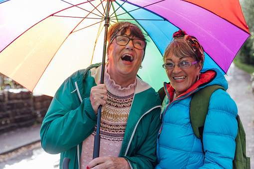 Waist up front view of two senior female friends on their way to begin their hike along the forest landscape surrounding Loch Torridon in the Northwest Highlands, Scotland. They are walking along a road wearing their waterproofs and using their umbrella to stay dry on a rainy day. They are laughing together.