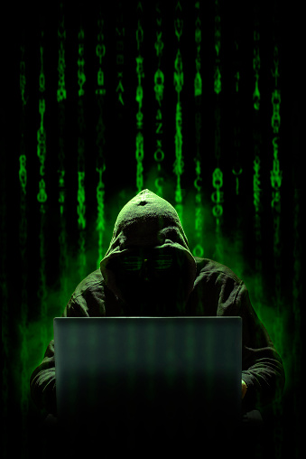 A hooded hacker works on a cyber attack with a laptop, against a background of matrix style computer code where a skull can be seen. Cybercrimes.