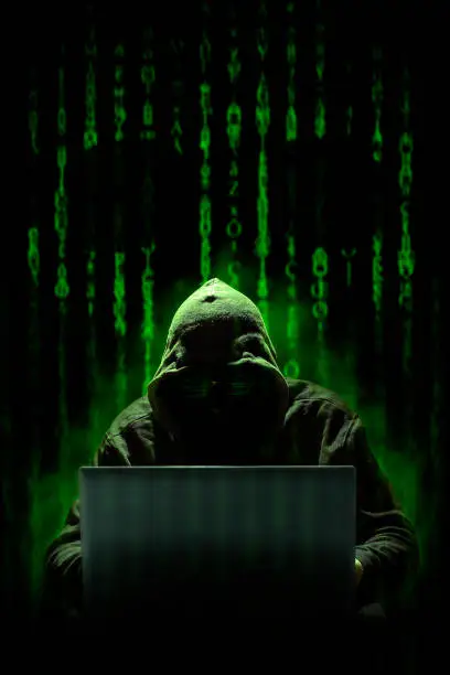Photo of A hooded hacker works on a cyber attack with a laptop, against a background of matrix style computer code where a skull can be seen. Cybercrimes.