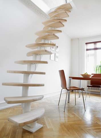 Spiraling staircase in white living room, with window and chairs around dining table. Staircase is specialmade to take up less room.