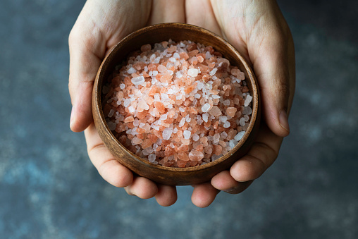 Hand is holding a bowl of Himalayan salt crystals to camera in front of dark blue background.