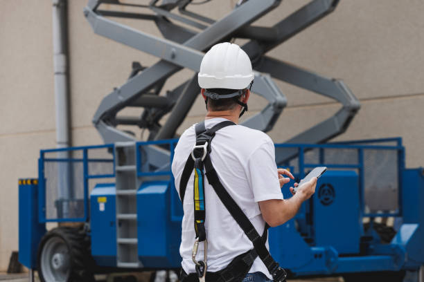 Engineer in a hard hat and harness with a smartphone looking an aerial work platform (AWP). stock photo