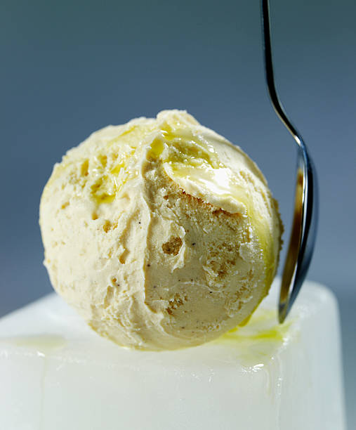 Ball of ice-cream on top of ice cube with spoon standing on the side, bluish grey background.