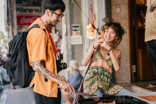 Asian woman on old town trip using smartphone to taking photos of boyfriend while he trying to grill a food.