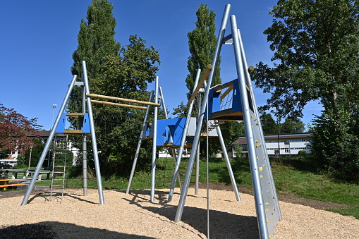 Playground for children with various equipment made of metal, wood and ropes. On the ground there is mulch because of safety reasons. There are trees on the background.
