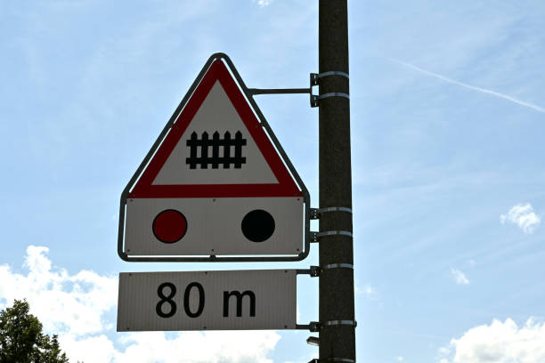 Three traffic signs arrange vertical are meaning railroad crossing ahead with barrier or gate with traffic light in distance of 80 metres. Triangular sign has red frame, white field and black barrier.