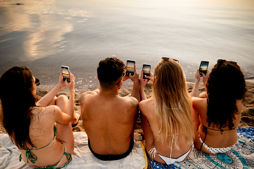 Rear view of group of friends sitting together on the beach taking photos of sunset with their mobile phones