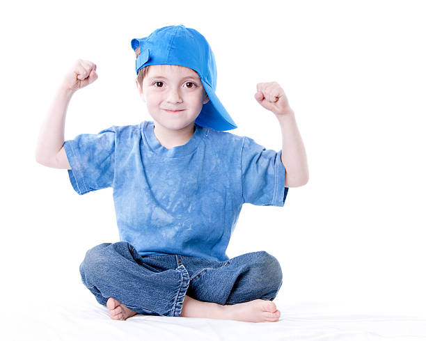 A full length image of a smiling caucasian little real boy sitting cross legged flexing his muscles, wearing a blue sh