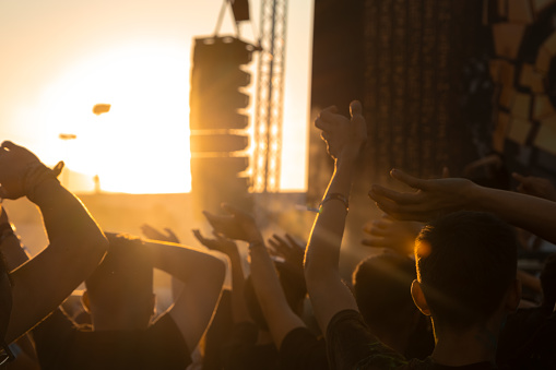 A photo of the crowd at a concert or music festival. The crowd is facing the stage, with their hands in the air. The photo is taken from behind the crowd, so you can see the backs of their heads and arms. The background consists of a stage with speakers and a large screen. The sky is visible in the background, and the sun is setting, creating a golden glow. The photo has a warm and energetic mood.